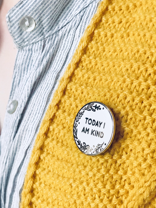 A  round white enamel pin with a floral design and the words "Today I Am Kind" is pinned to a yellow cardigan.