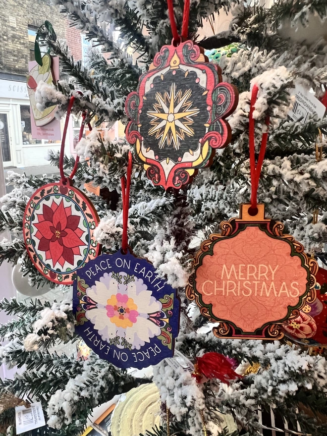 A set of four multi coloured wooden tree decorations are hanging by red ribbons on a fake white christmas tree with other decorations. 