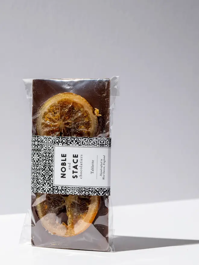 Orange Confit Chocolate Tablette wrapped in clear plastic, standing on a white table. 