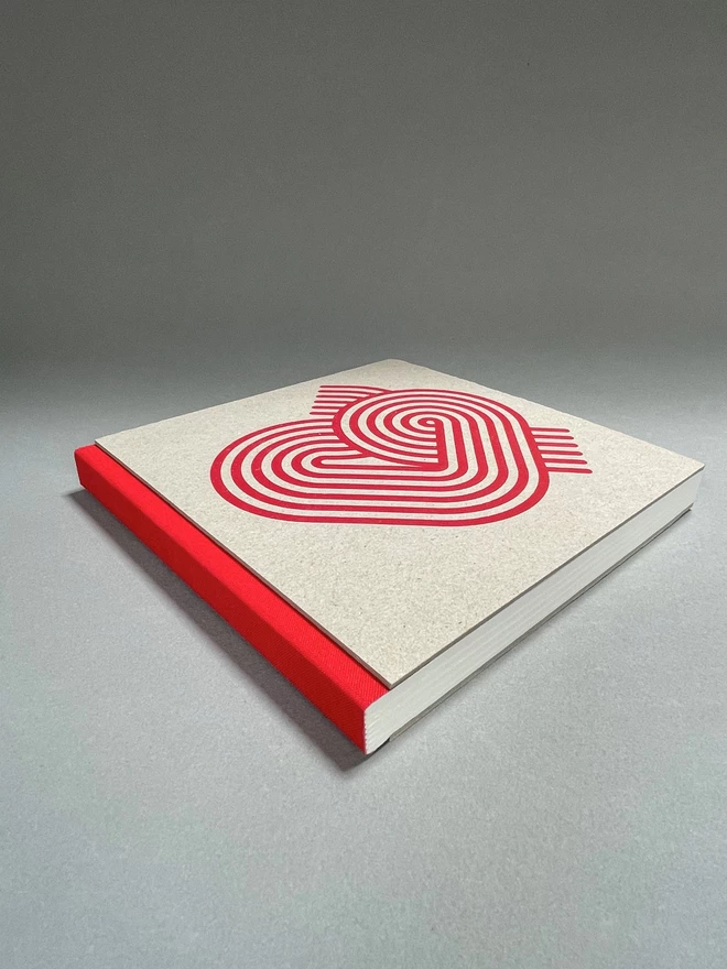  A pasteboard sketchbook with a red spine and a stripy heart design, lies on a light grey studio backdrop. Red fabric spine towards us.