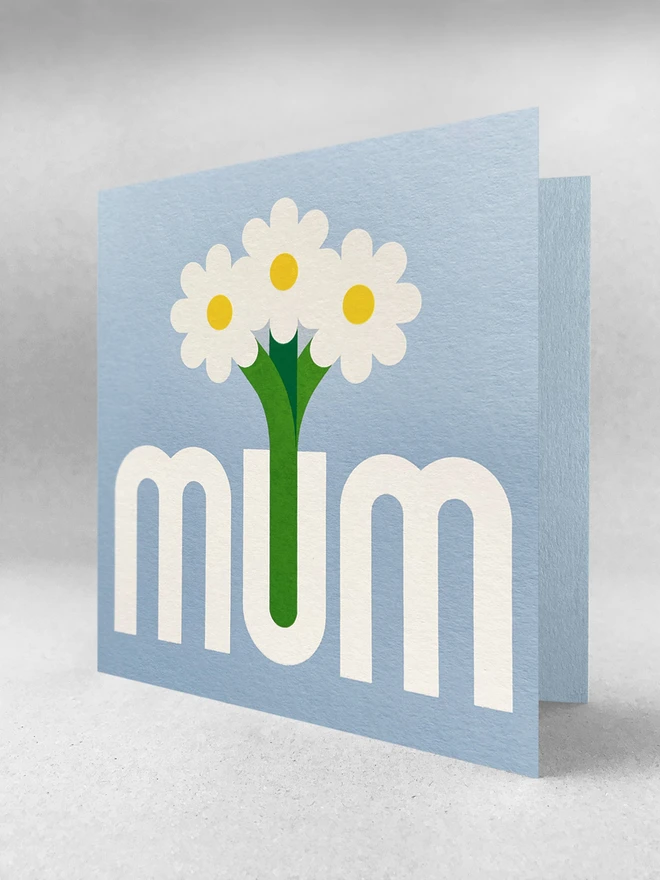 light blue square card with mum printed in grey, the U is a vase with three flowers stood in it. Card is stood in a light grey space with soft grey shadows.