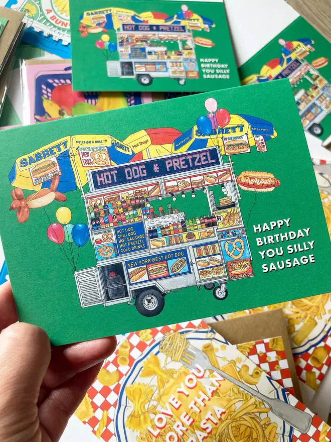 Food-inspired illustrated greeting card featuring a vibrant New York Hot Dog food truck, surrounded by birthday balloons, set against a bold green background. A fun, colourful design perfect for food lovers and anyone celebrating a birthday