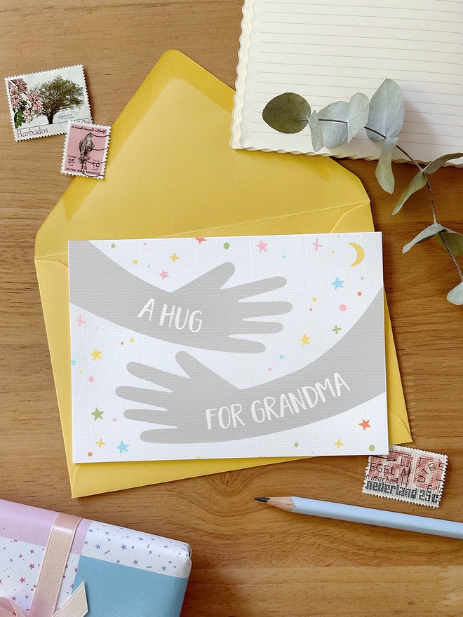 A friendship greetings card with a hugging arms design and personalised wording that reads "A hug from..." lays on a yellow envelope on a wooden desk.