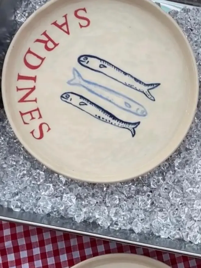 'Sardines' Red Wording Ceramic Side Plate With 3 Fish, a ceramic side plate sitting on ice with a fish design. 