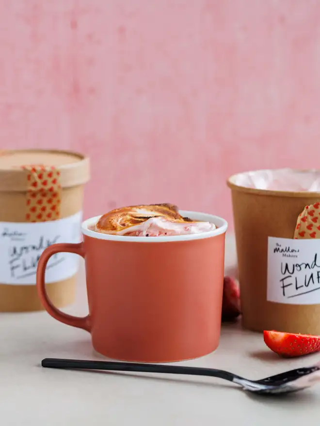 Strawberry Wonder Fluff Tub, a hot chocolate topped with torched strawberry fluff, in front of one open and one sealed tub. This is in front of a pink background with fresh strawberries scattered in front.