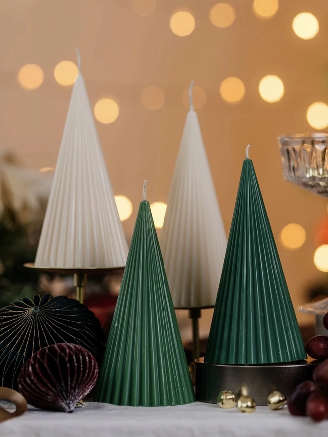Green Christmas Tree Cone Candles. Ribbed cone candles in forest green and cream, styled on gold stands with velvet and paper baubles, gold jingle bells and red grapes against warm bokeh lights. The mix of colours makes a striking, modern Christmas centrepiece.