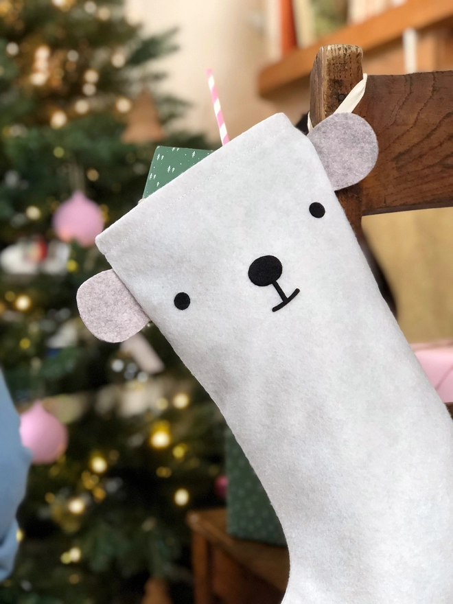 A handmade felt polar bear stocking hangs on a wooden chair, where a little girl sits, in front of a Christmas Tree.