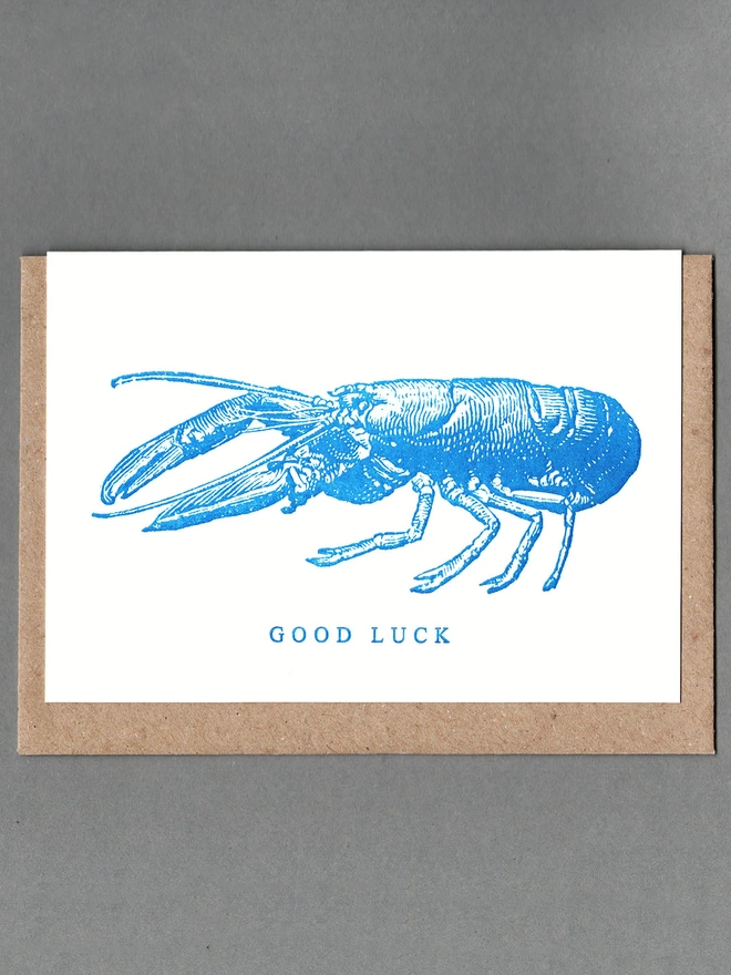 White card with blue illustration of a lobster and text reading 'GOOD LUCK' with a brown envelope behind