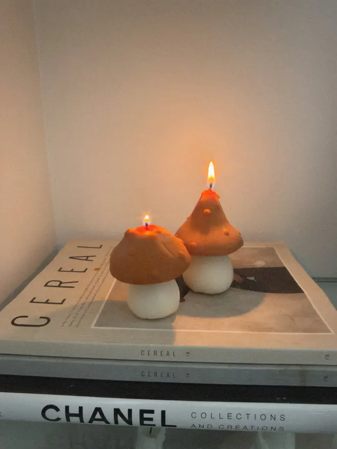 Pair Of Little Mushroom Candles (Set Of 2). Two burning mushroom candles with orange dotted caps and cream stems, placed on a stack of books against a softly lit wall. The cute sculpted candles make a cosy, giftable piece for candle lovers.