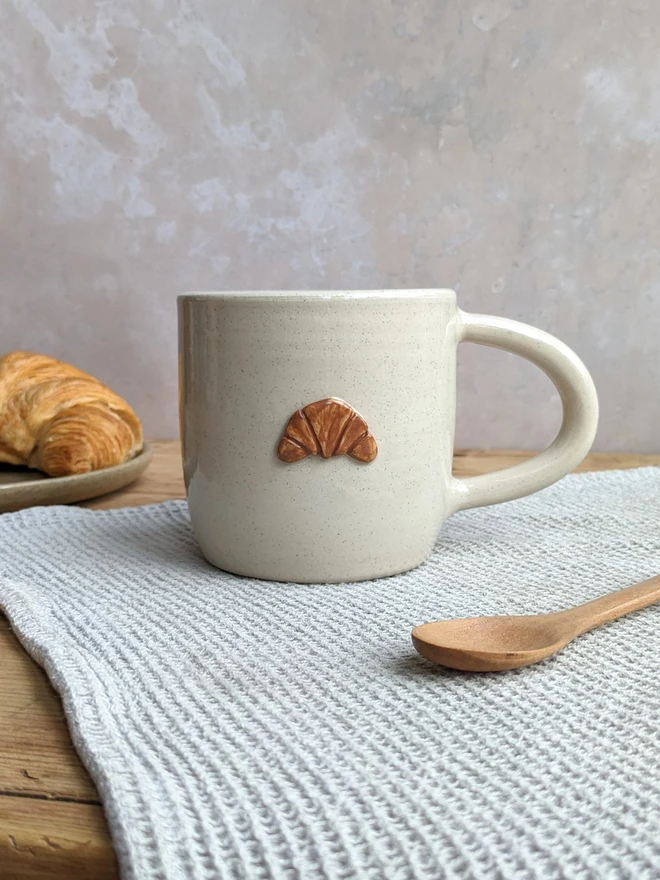 A large white ceramic mug affixed with a croissant on the front of the mug. The mug sits on a wooden table.