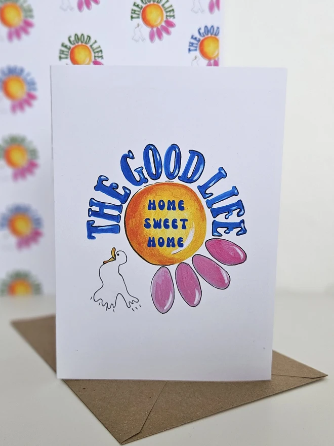 A white card with an illustration of a pink flower and a white bird. The Good Life is written using a retro font style in blue. In the centre of the flower it says Home Sweet Home. 