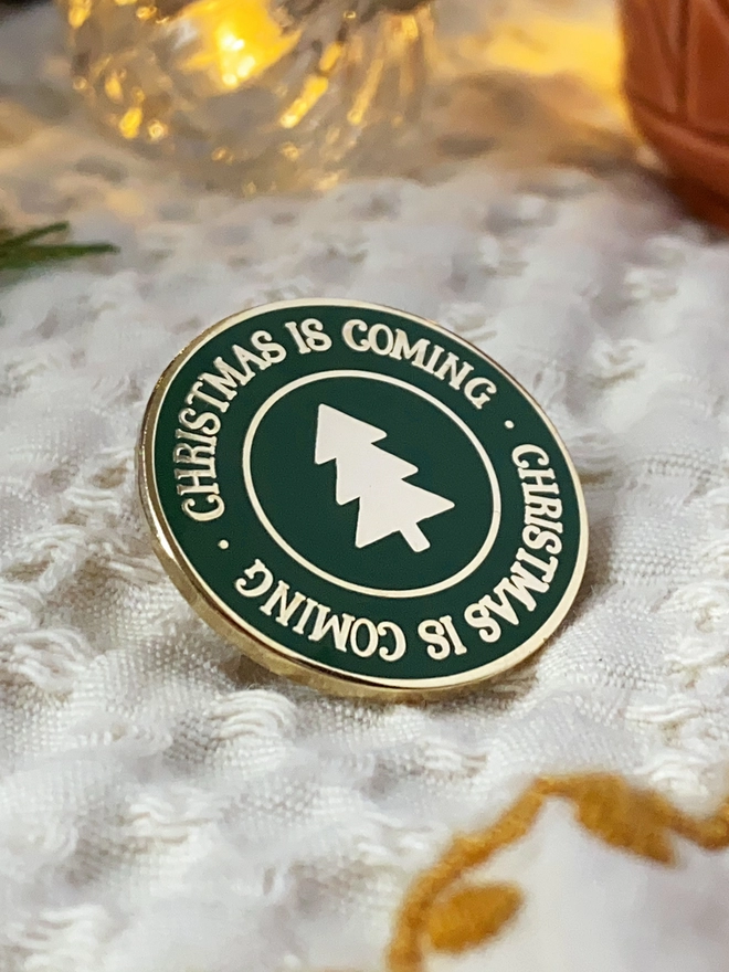 A deep green round enamel pin badge with a gold Christmas tree in the centre and gold lettering around the outside that reads "Christmas Is Coming" rests an ivory fabric.