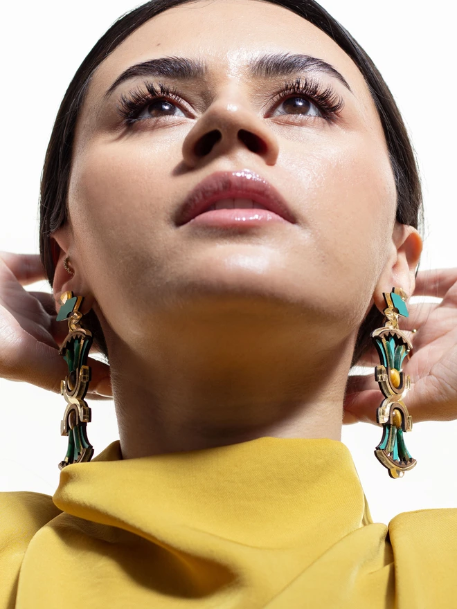 A person wearing gold and green earrings with a blurred background