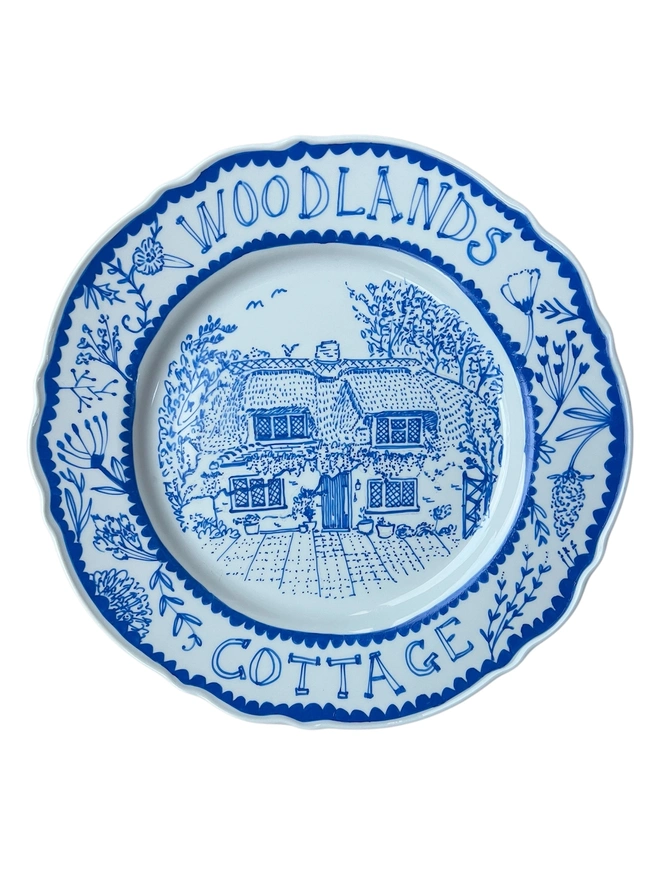 Medium dinner plate illustrated in a fun whimsy style, with the name of the cottage over the top of the plate rim, a botanical border around the rest, and a cute cottage inset, all painted in blue ink on white plate.