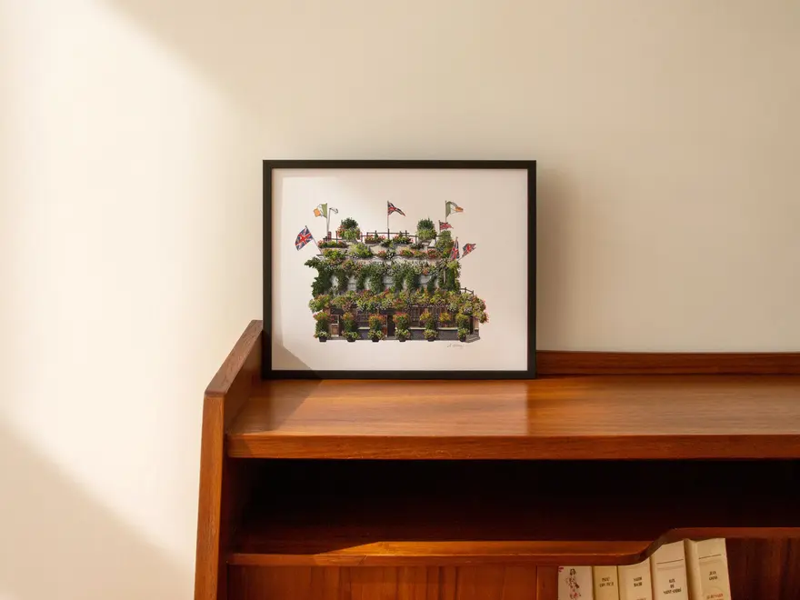 Kensington Churchill Arms Giclée Print (Unframed), a print of a pub in a black frame against a wooden surface sitting on a plain backdrop. 