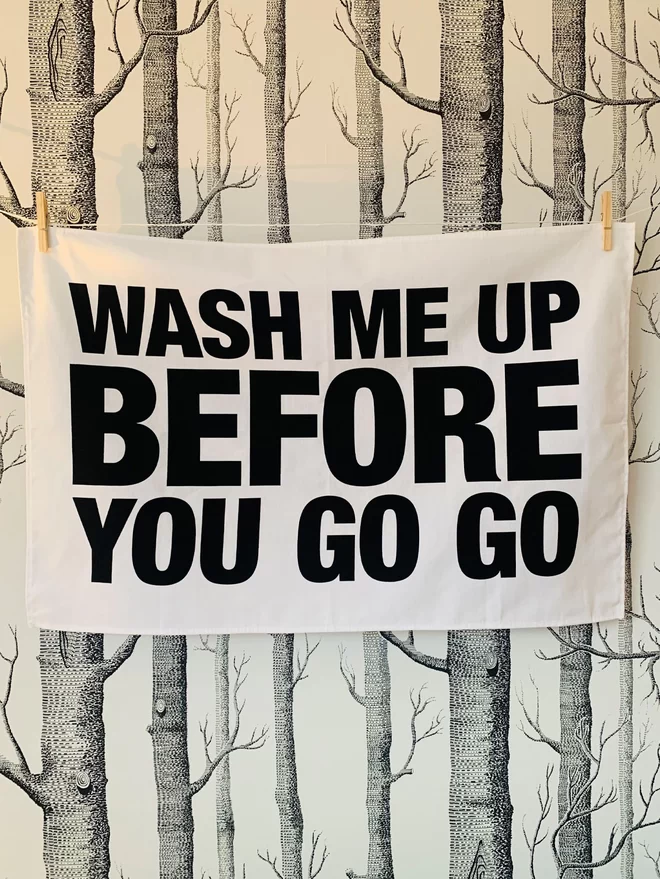 London Drying Wash Me Up Before You Go Go black text on white tea towel hung washing line style in front of trees/woods black on white patterned wallpaper