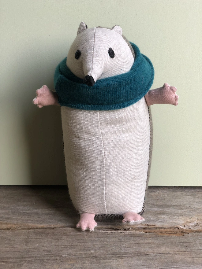 Handmade fabric toy hedgehog made from natural linen (front) and brown tweed (back). Wearing a blue lambs wool scarf. Hands out stretched and embroidered eyes, nose and smile