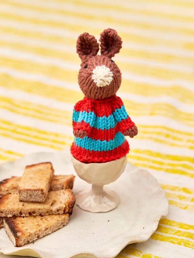 Egg Cosy Rabbits Easter Table Decoration, a colourful Easter cosy rabbit sitting on a striped tablecloth surface. 