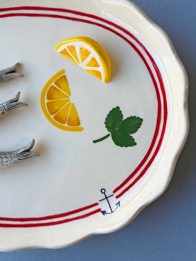 ceramic white platter with red border and anchor detail, with 3 ceramic sardines arranged on top, a ceramic wedge of lemon plus lemon painted on, with parsley detail, on a blue background