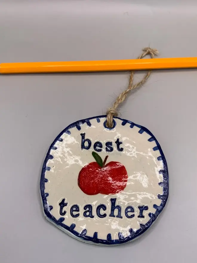 'Best Teacher' Handcrafted Ceramic Ornament, a ceramic apple ornament sitting on a plain white surface hanging from a pencil. 