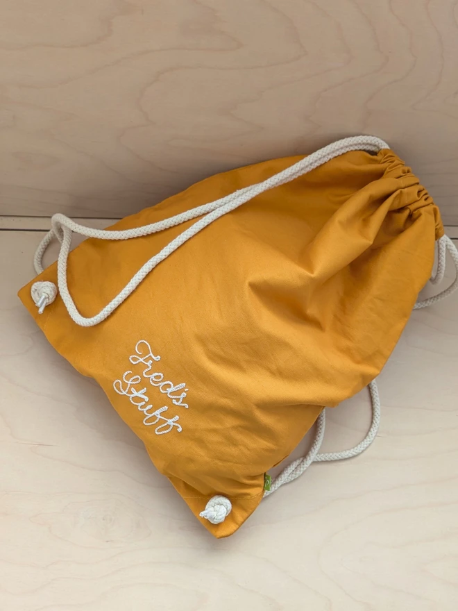 Mustard Gymsac Drawstring Bag casually sitting on a wooden bench, reading 'Fred's Stuff' in White embroidered cursive lettering