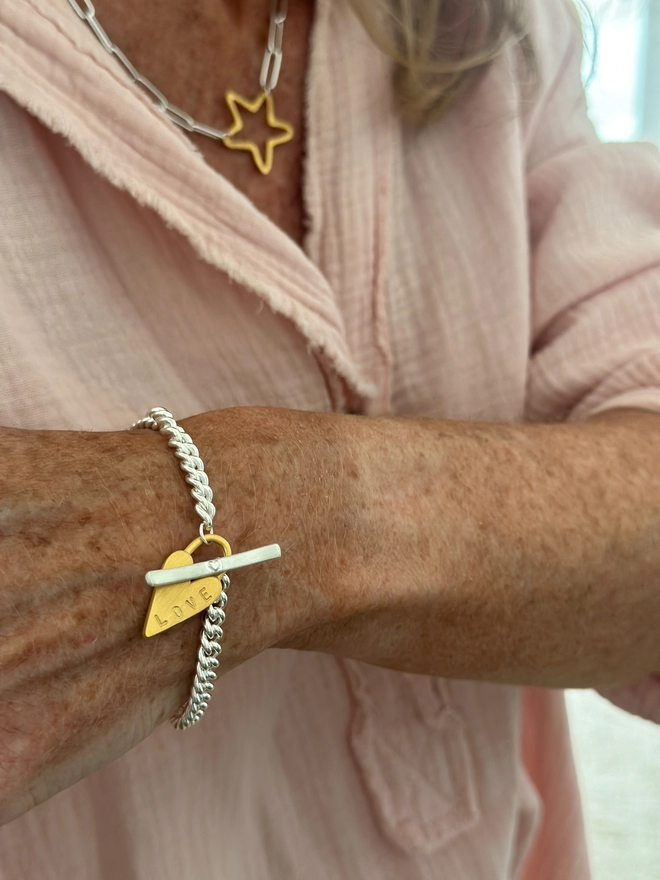 model wears a sterling silver chunky curb bracelet with silver t bar fastener and personalised gold chunky heart padlock charm