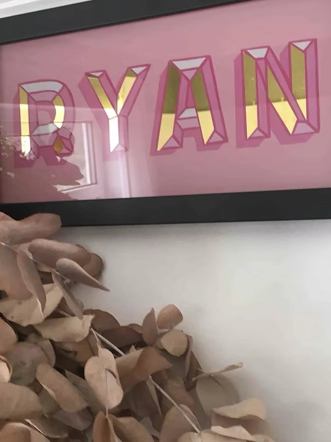 Personalised Name Wall Plaques, a pink personalised gold leaf sign sitting in a black frame hanging in front of a white wall. 
