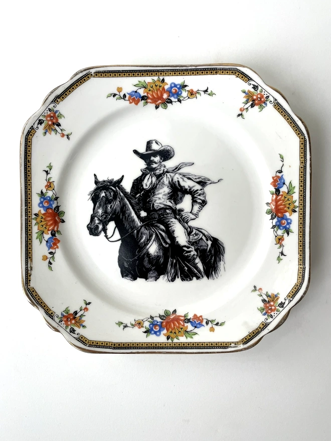 vintage plate hanging on a white wall, it has a decorative border, with a printed vintage illustration of a cowboy in the middle 