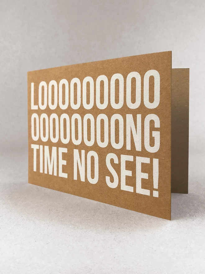Looooooong time no see screenprinted across this card with multiple o’s in the Long, to emphasise the LONG. Printed in white ink on a brown kraft card stock in landscape format. Stood slightly open, in a light grey studio set.
