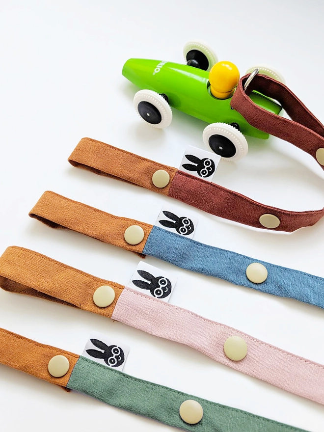 Handmade toy straps in woodland shades, featuring sturdy snap fastenings and small fabric tags with a bunny logo, neatly arranged in a row.