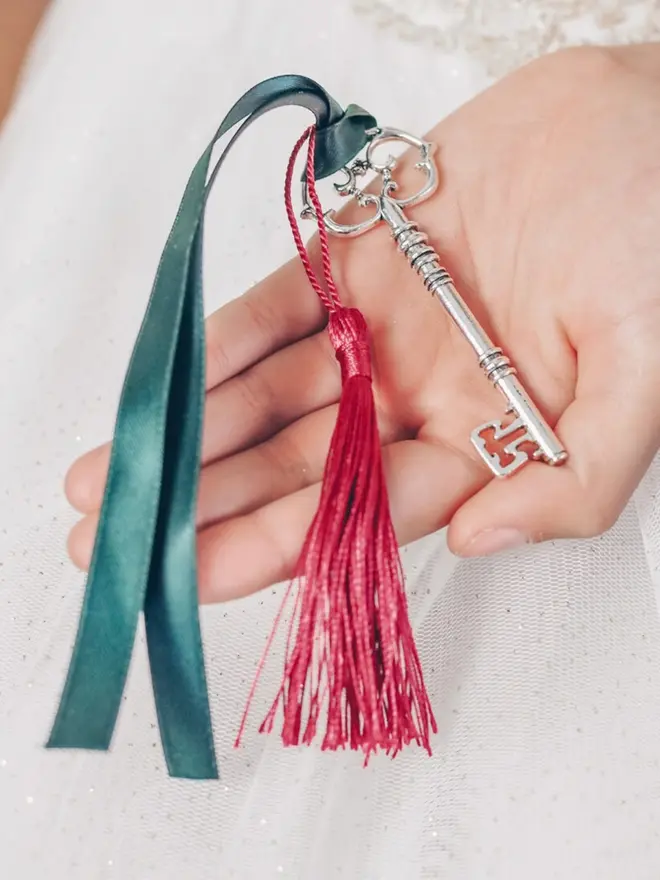 An ornate silver key rests on a child's open palm. A green satin ribbon and red silk tassel are tied to its bow.