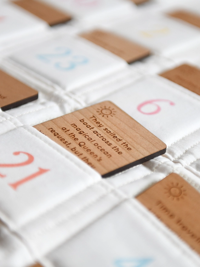 An ivory fabric advent calendar with 24 numbered pockets contains wooden cards with story ideas engraved on them. One card pokes out of the number 21 pocket.