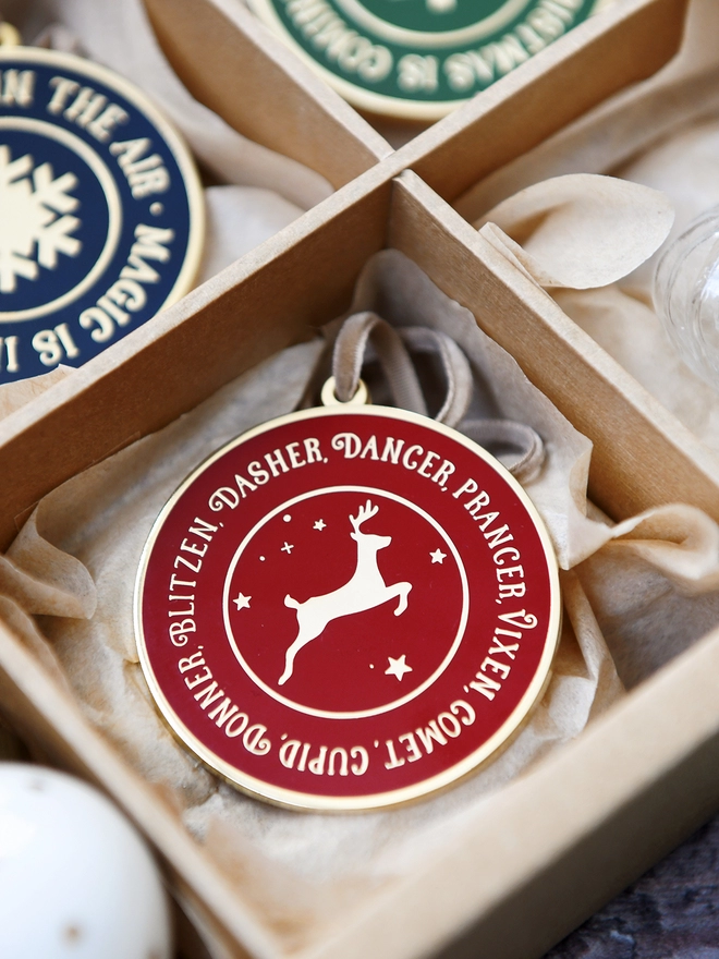 A deep red and gold enamel Christmas decoration, with the names of all eight reindeer surrounding a gold reindeer, is tucked into a sectioned box.