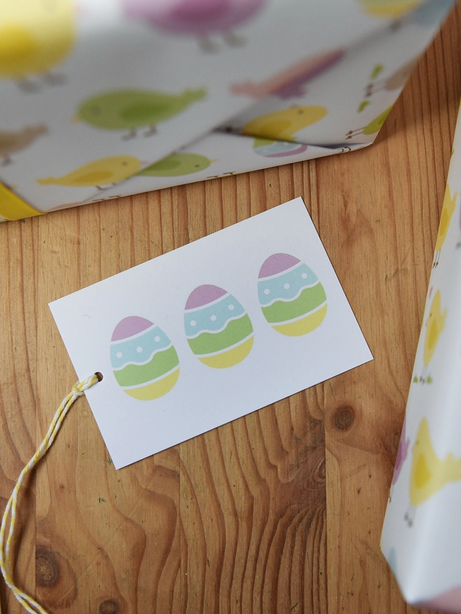 A gift wrapped in pastel spring chicks wrapping paper tied with yellow ribbon has a gift tag with three easter eggs on it.