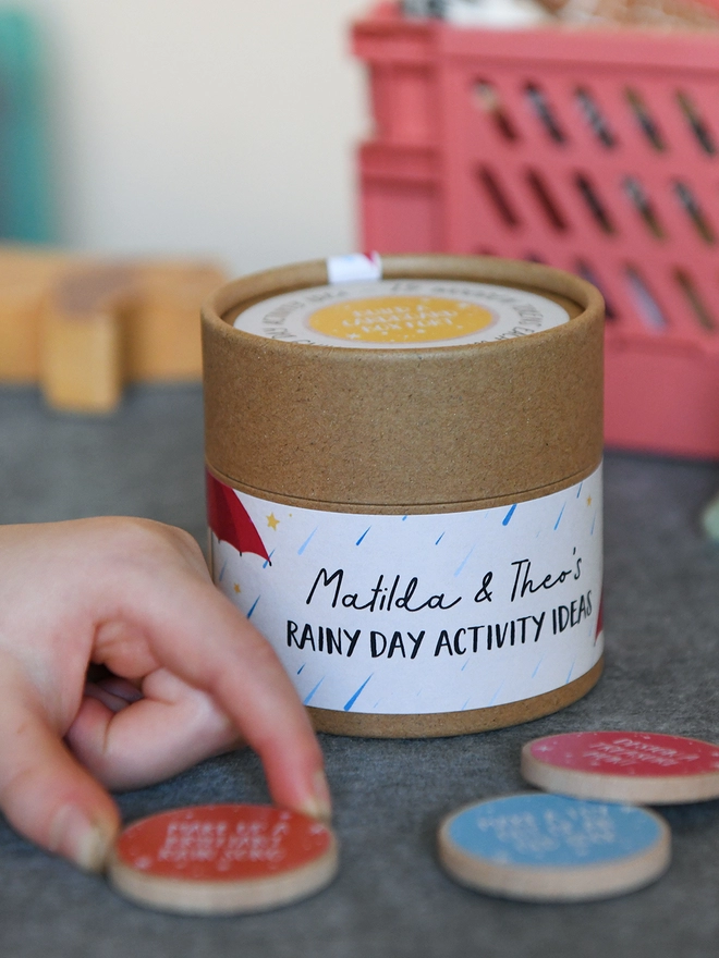 A toddler holds a cardboard jar with a label that reads Rainy Day Activity Ideas. There are several wooden tokens around the jar.