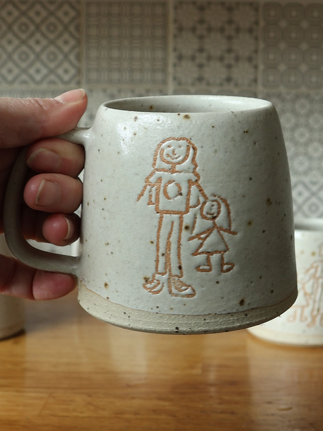 Childrens Drawing Handmade Ceramic Mug held in a hand over a wooden table. The mug features a child's drawing of a mother and child