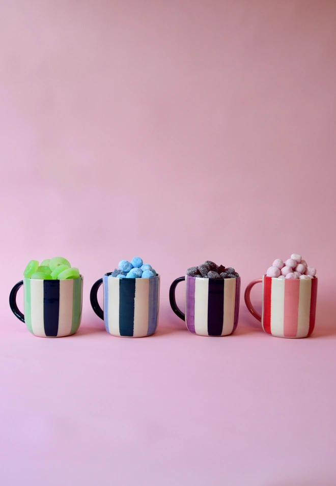 Handmade striped ceramic mugs in four colourways – green, blue, purple and pink – filled with sweets and styled against a soft pink background.