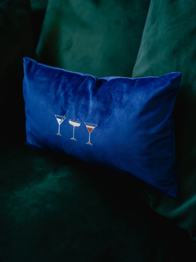 A navy blue cushion with cocktail embroidery on a green sofa