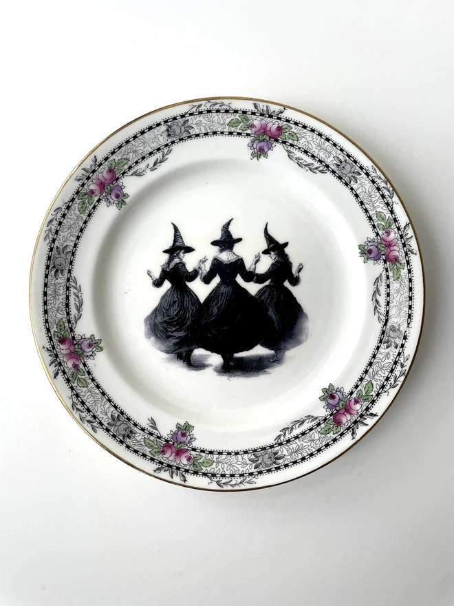 a coven of 3 witches dance hand in hand, the image is victorian in black and white and is printed directly onto an ornate vintage plate
