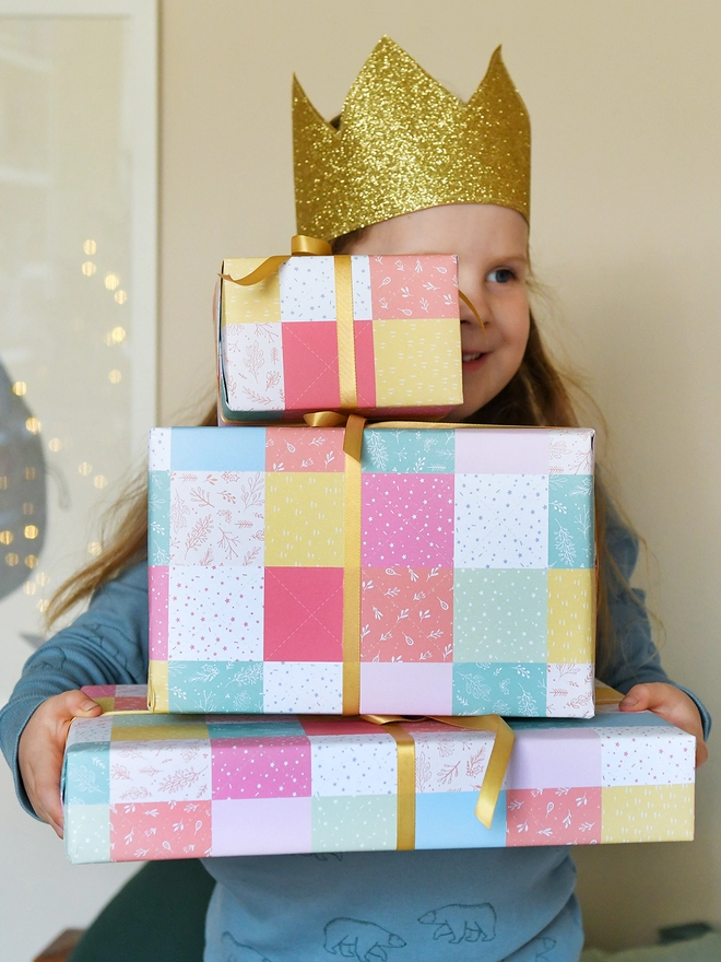 A young child wearing a gold crown is holding a pile of gifts wrapped in gift wrap with a design of pastel patchwork quilt design.