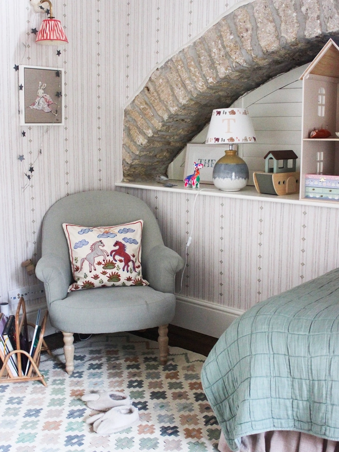 Cosy children’s bedroom featuring a personalised initial “T” table lampshade with woodland animal motifs, styled beneath a rustic stone arch. The nursery interior includes a soft green armchair, patterned rug, wooden toys, doll’s house and sage green bedding.