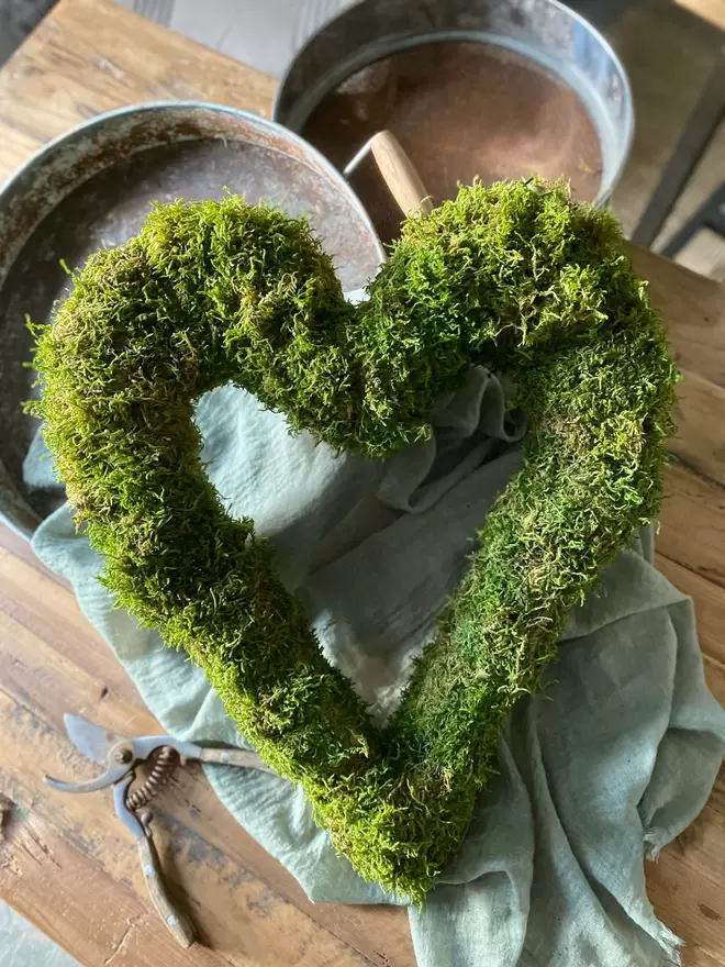 A hand bound moss heart wreath lays against a silver bucket, a duck egg blue cloth drapes over the bucket. 