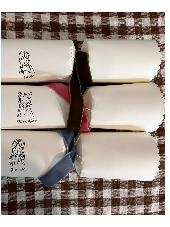 three white christmas crackers, each with a bespoke illustration of a child or pet, with different coloured velvet ribbon. sitting on brown and white gingham table cloth