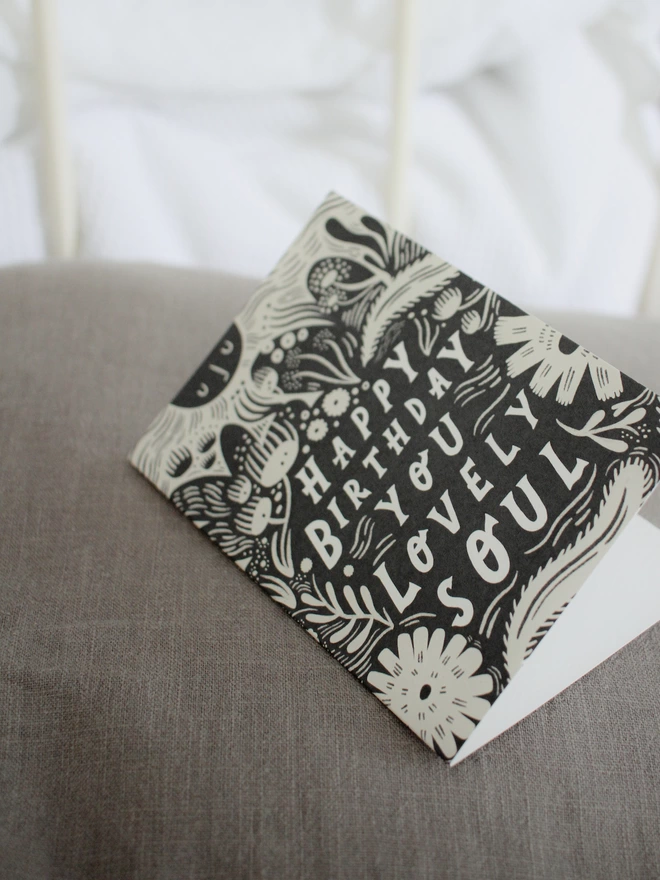 Black and white greeting card with illustration and the words Happy Birthday you lovely soul written on it lying on top of a grey cushion