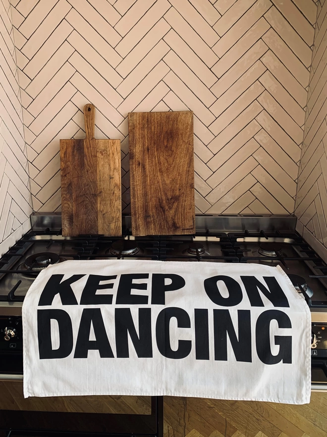 London Drying Keep On Dancing black screen printed text on white tea towel laying on kitchen/cooker with 2 timber chopping boards leaning against tiled wall in background