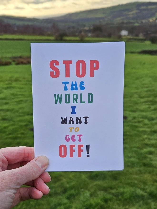 a white card with colourful writing that says stop the world I want to get off in different font styles held by a hand against a backdrop of the countryside