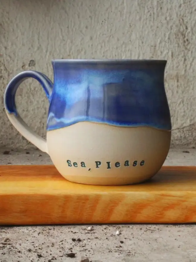 A 'Sea, please' ceramic mug with a blue glazed top and unglazed ceramic bottom resting on a wooden surface.