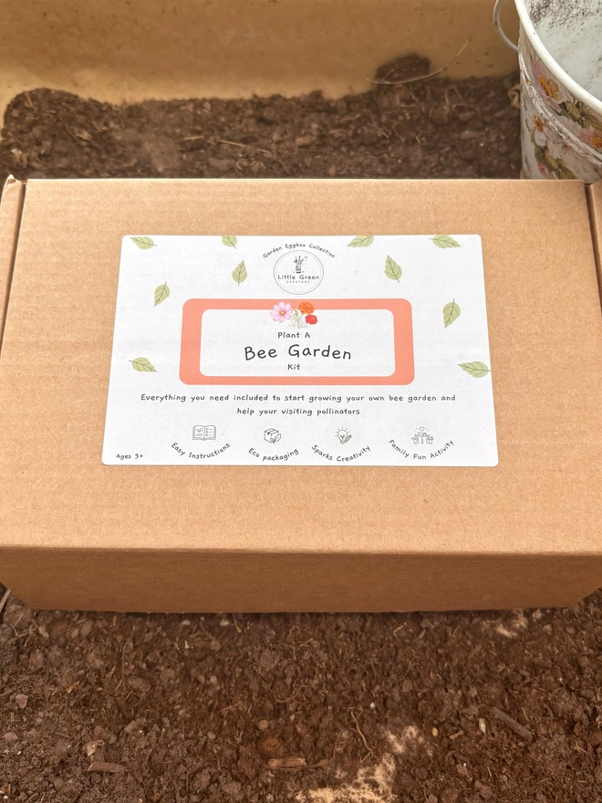 Cardboard box with “Plant a Bee Garden” label on. Placed on soil with a white floral bucket in the background.