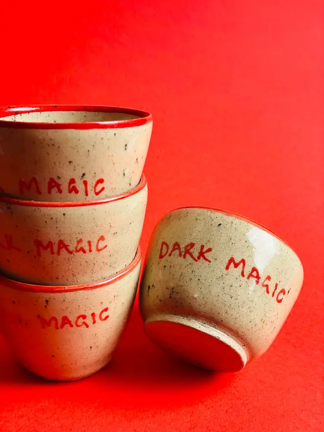 'Dark Magic' Mug, a group of ceramic mugs stacked together in front of a red background. Each mug says 'Dark Magic' and is stamped with a signature red dot on the bottom. 