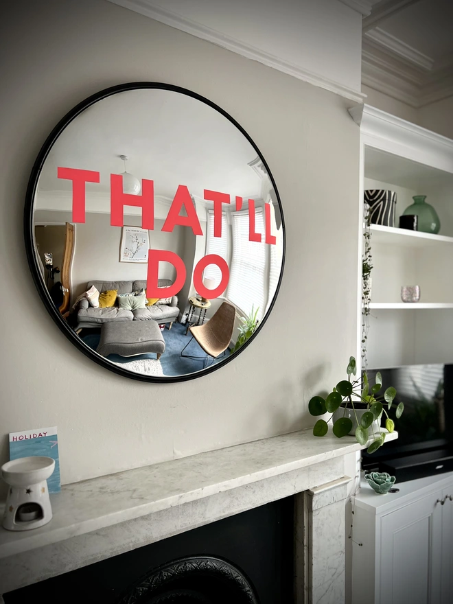 Round convex mirror with 'That'll Do' handpainted in neon pink above a white fireplace with plants